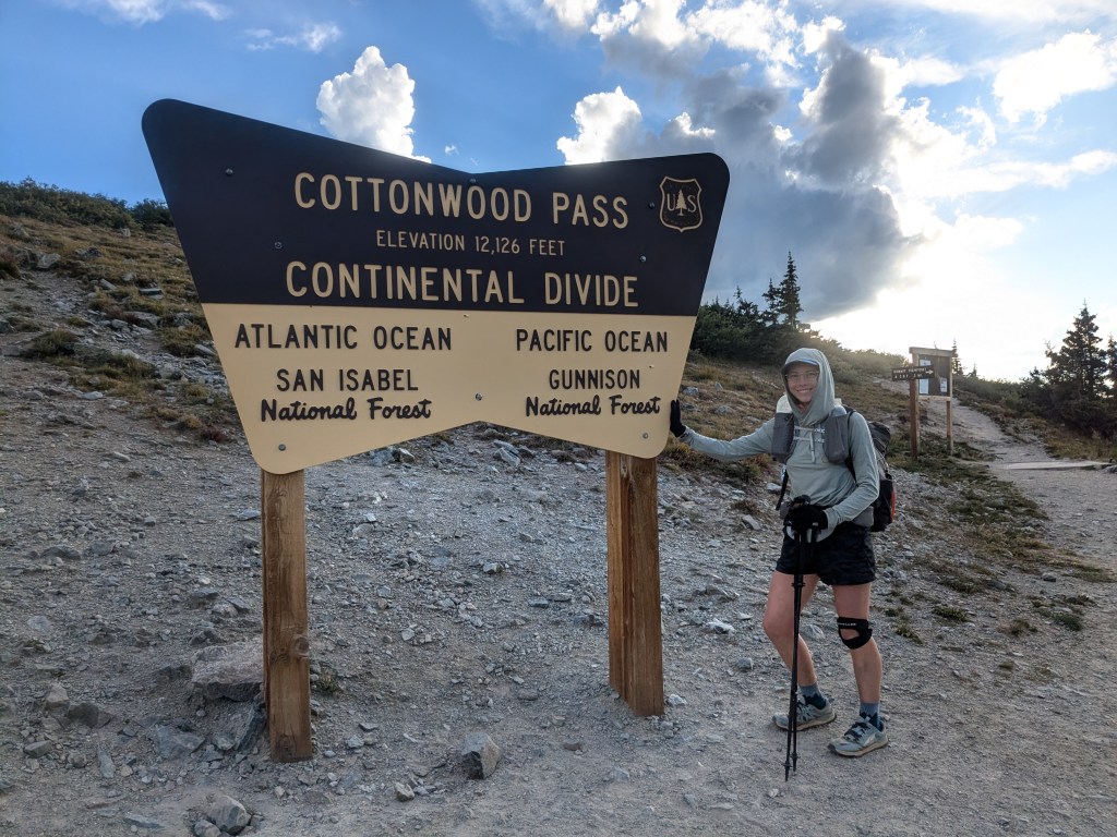 A hiker standing next to a sign that says "Cottonwood Pass elevation 12,126 feet Continental Divide"