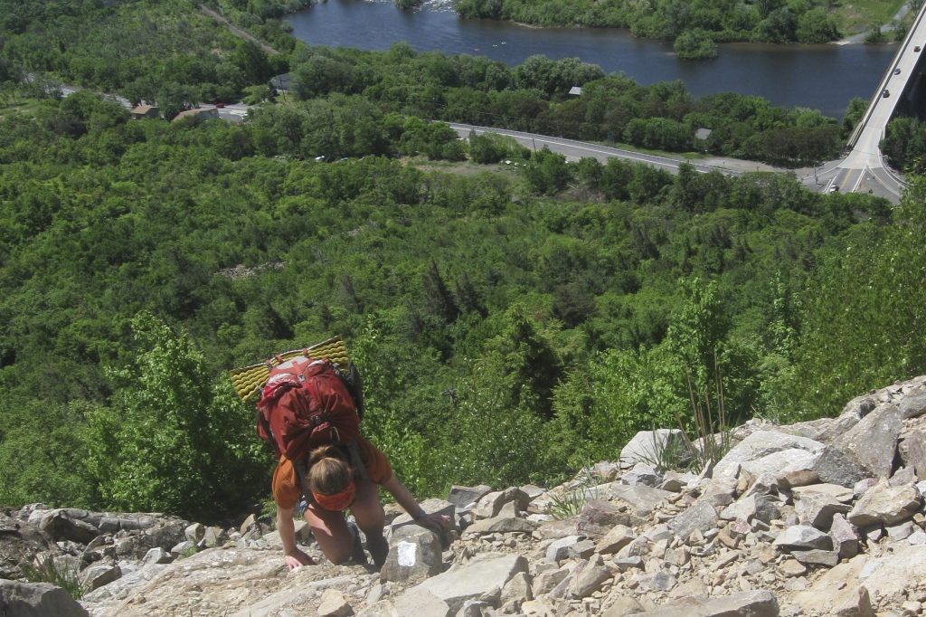 Backpacker using her hands to climb up a very steep trail above a road