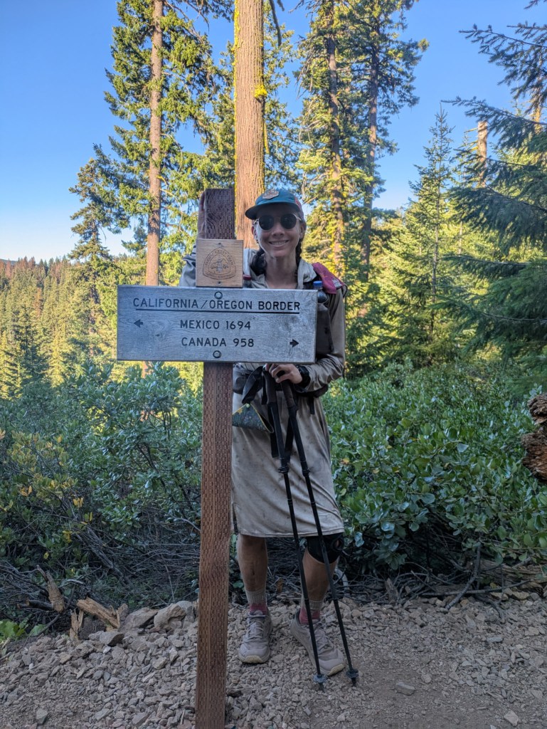 hiker standing behind a sign that says "California/Oregon Border" and "Mexico 1694" and "Canada 958"